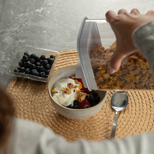 moça segurando pote drop e despejando cereal em uma bandeja com frutas e iogurte